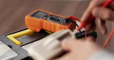  person checks a battery's voltage using an electronic multimeter in a well-lit workspace. 