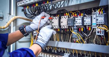  A person in blue overalls is repairing electrical equipment, focused on their task in a workshop setting.  